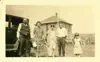 Image shows a black and white photo of a group of people with a house in the background and an automobile to the left. 