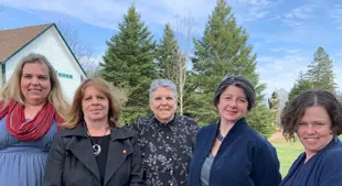 Image shows five women standing shoulder-to-shoulder with coniferous trees in the background. 