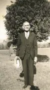 Image shows a black and white photo of a man in a three-piece standing outdoors and holding a hat. 