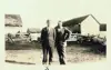 Image shows a black and white photo of two men standing in front of farm buildings. 