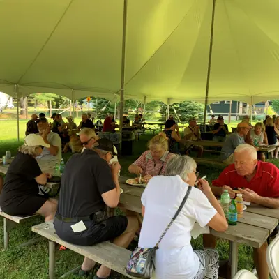 Image shows people eating at picnic tables in a large tent. 