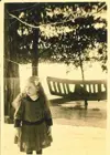 Image shows a black and white photograph girl standing in front of trees with a lake in the background. 