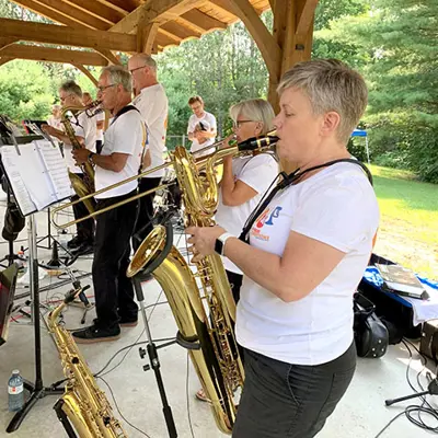 Image shows a band playing music with a woman playing a saxophone in the foreground. 