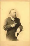 Image shows a black and white portrait of a well-dressed man holding a book. 