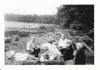 Image shows a black and white photo of a group of people eating lunch on a beach strewn with logs. 