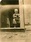 Image shows a black and white photo of a child in the corner of a doorway. 