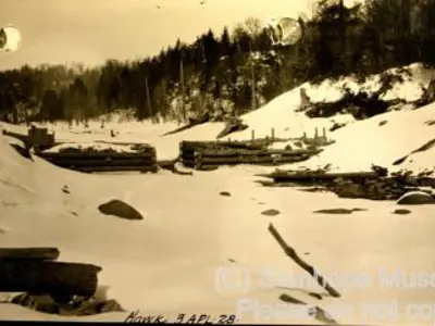 Image shows a black and white photo of the log chute under construction in winter. 