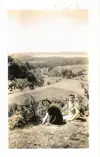 Image shows a black and white photograph of two women seated on the edge of a cliff with field and forest below and a lake in the background. 