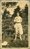 Image shows a black and white photograph of a woman standing in front of a garden. 