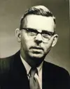 Image shows a portrait of a man with horn-rimmed glasses in suit and tie. 