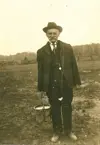 Image shows a black and white photograph of a man wearing a three-piece suit and hat standing outdoors. 